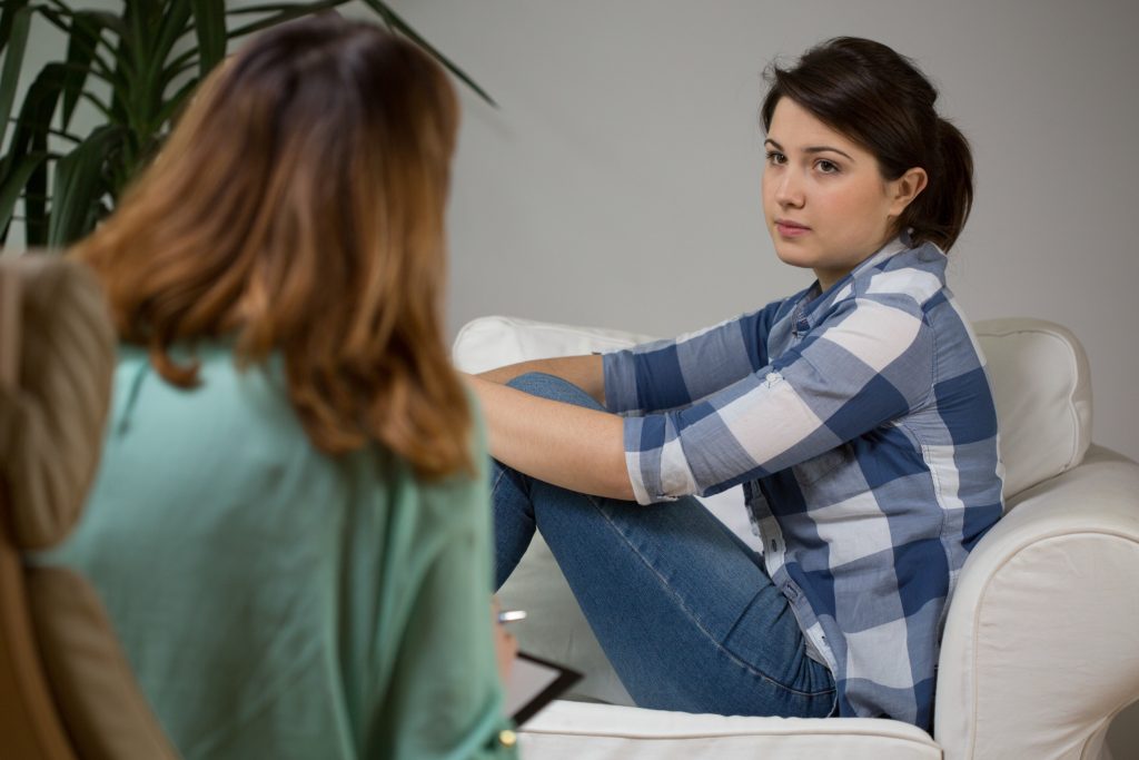 Image of a teen girl, sitting on a couch, holding her knees, while speaking with a teen therapist. Learn how a skilled teen therapist in Englewood, CO can help your teen cope with trauma, anxiety, and more.