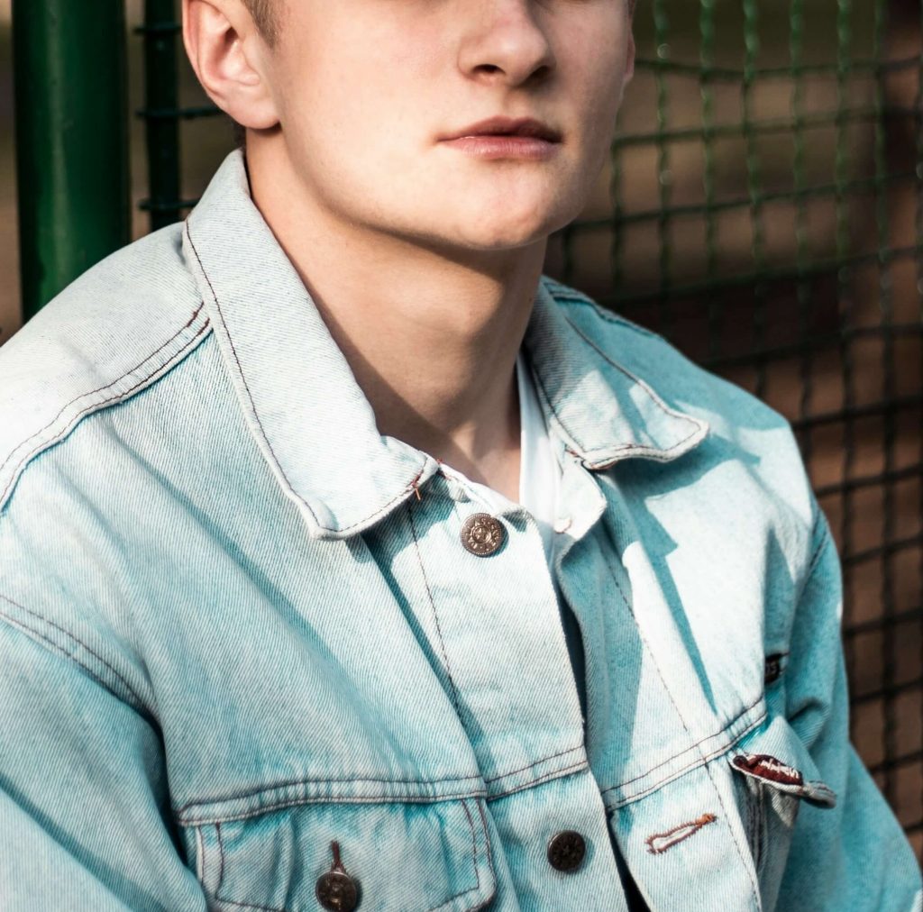 Image of a teen boy, wearing a jean jacket, leaning against a fence. Help your teen learn to manage their emotions with the help of a teen therapist in Denver, CO.