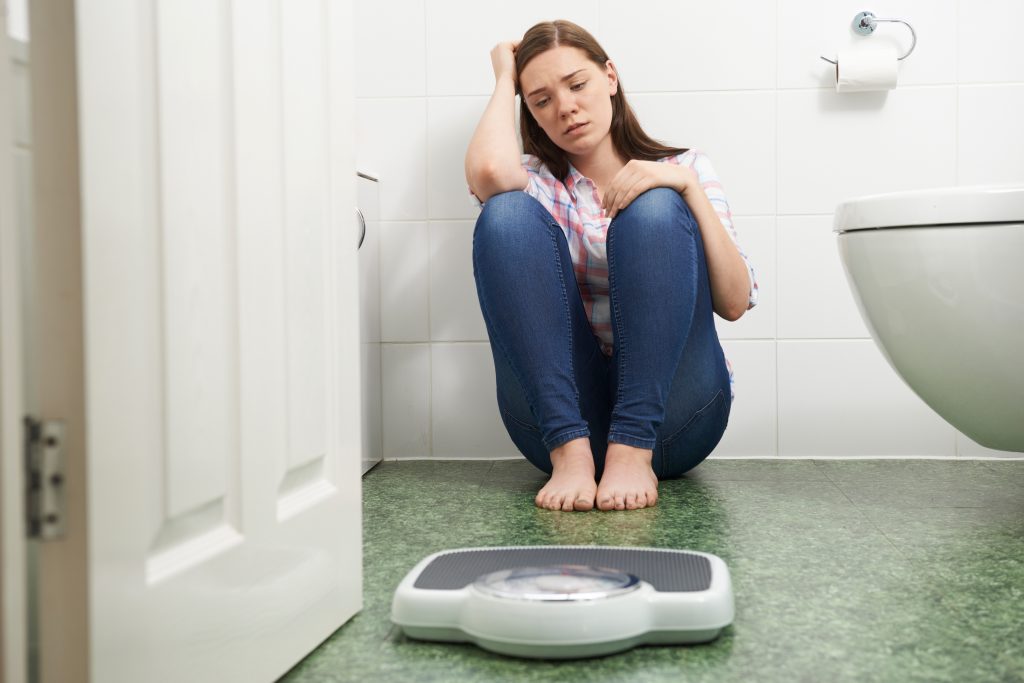 Unhappy Teenage Girl Sitting On Floor Looking At Bathroom Scales