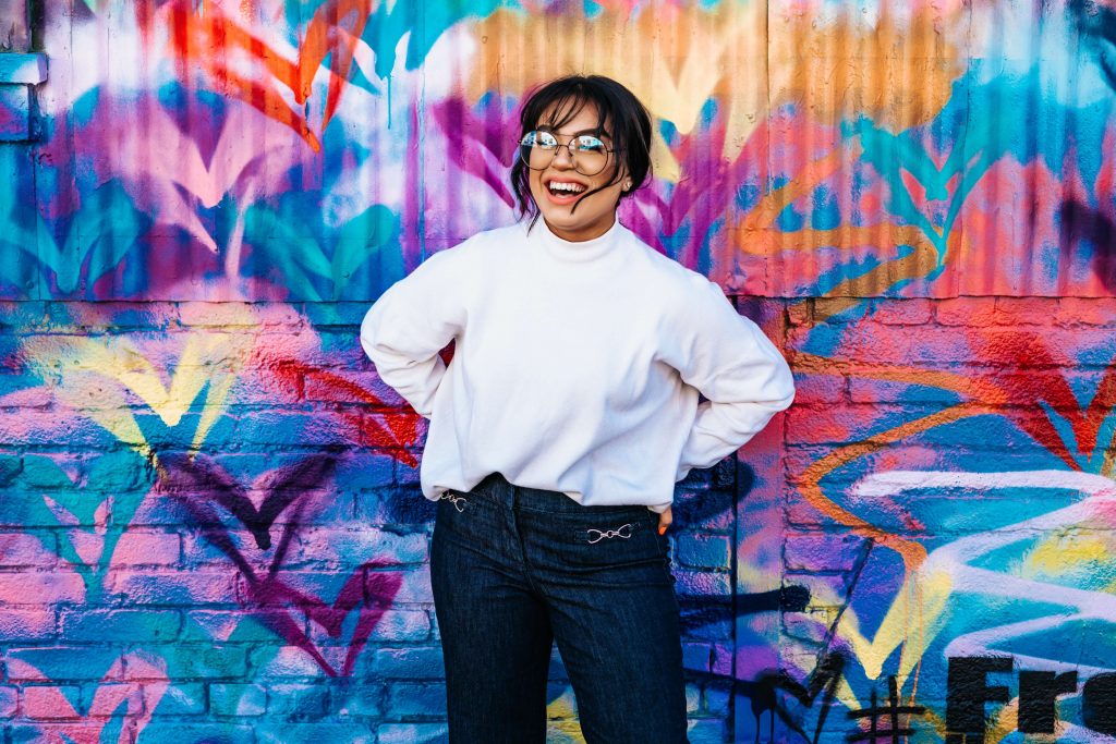 Image of a smiling woman wearing glasses standing in front of a colorful mural. Learn how effective EMDR therapy in Denver, CO can be when it comes to managing your trauma symptoms.