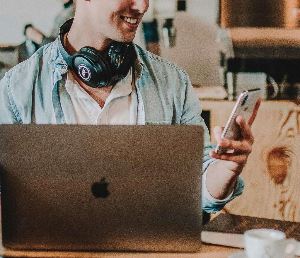 Image of a young man, sitting in a coffee shop at a table, smiling, while wearing headphones around his neck and holding a phone. Discover how EMDR therapy can help you cope with past trauma with telehealth in Colorado.