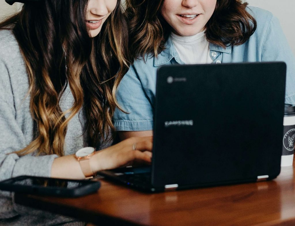 Image of two teen girls smiling and looking at a laptop. Discover the benefits of teen counseling via telehealth therapy in Colorado.