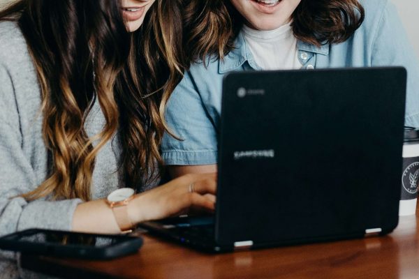Image of two teen girls smiling and looking at a laptop. Discover the benefits of teen counseling via telehealth therapy in Colorado.