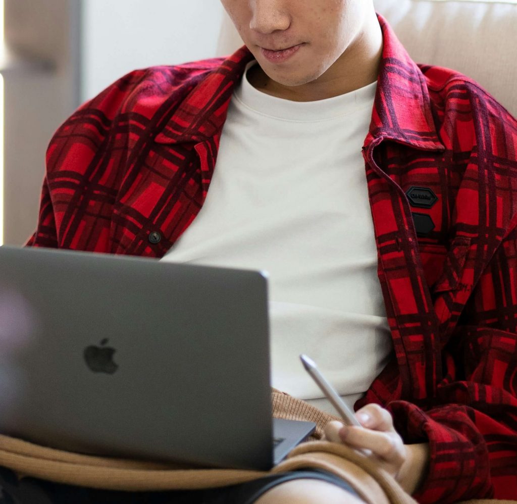 Image of a young teen boy wearing a red plaid shirt sitting on a couch holding a laptop. Help your teen find support with the help of teen counseling via telehealth therapy in Colorado.