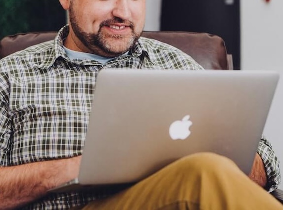 Image of an middle aged man sitting in a recliner, smiling and using a laptop. Looking for effective ways to cope with anxiety, trauma, and more? Learn how telehealth therapy in Colorado can help!
