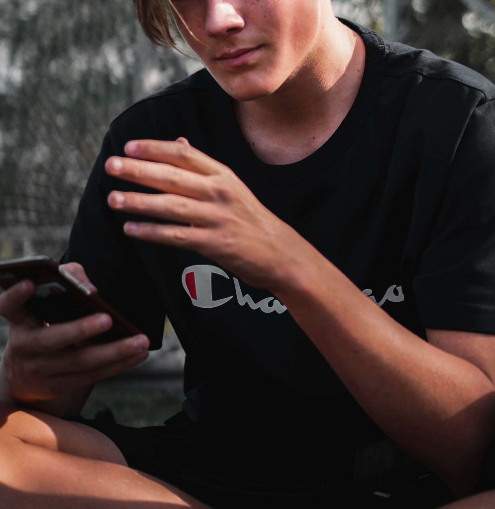 Image of a teen boy sitting on the ground outside holding a cell phone. Learn the benefits of teen counseling in Littleton, CO and how it can help your teen!