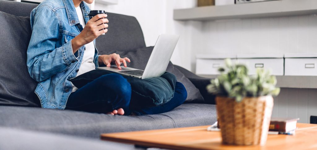 Image of a woman sitting on a couch holding a cup and laptop. Begin telehealth in Denver, CO to overcome your anxiety and more in convenient ways.