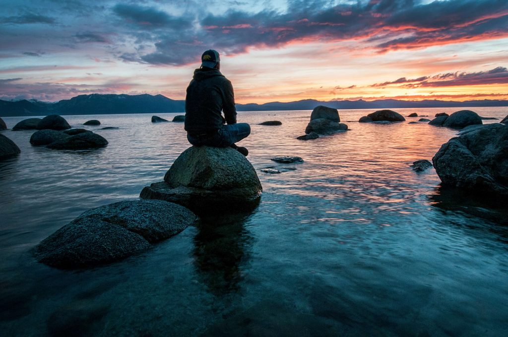 Image of a man sitting on a rock in a lake looking at the sunrise. Meet with a neurofeedback therapist to start managing your anxiety symptoms. Learn how neurofeedback for anxiety can help you!