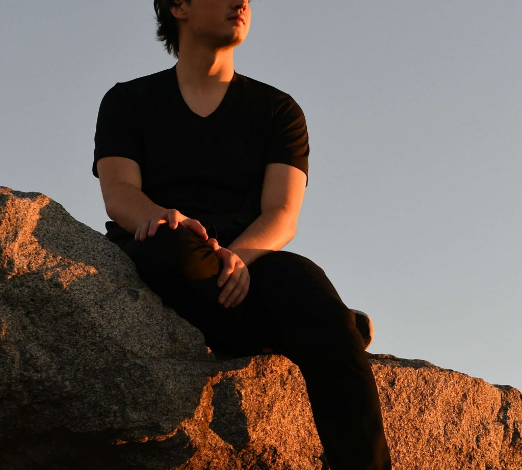 Image of a young man wearing all black sitting on a rock while the sun shines on him as it sets. Learn to manage your ADHD symptoms with the help of neurofeedback therapy in Denver, CO.