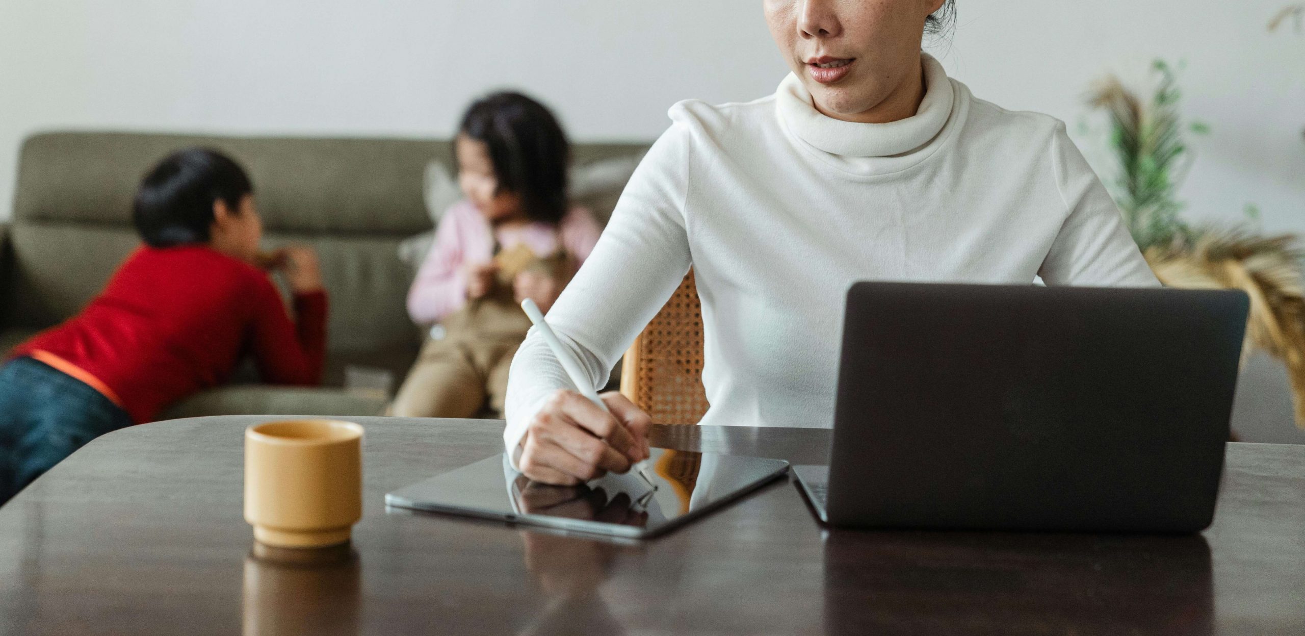 A busy mom works on her tablet during online therapy in Colorado while her children play in the background, highlighting therapy for moms and online therapy that takes insurance. 